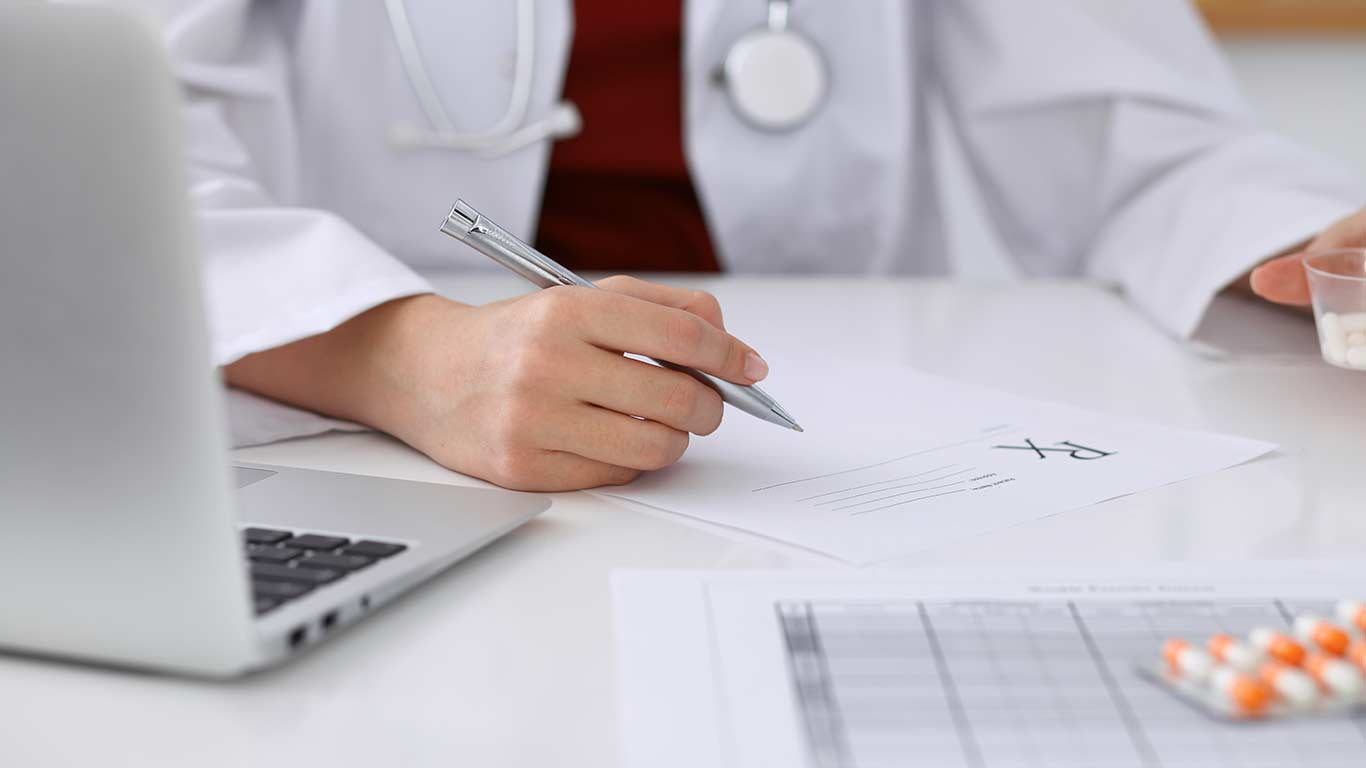 Closup of doctor's hands while filling out papers, laptop, and pills 